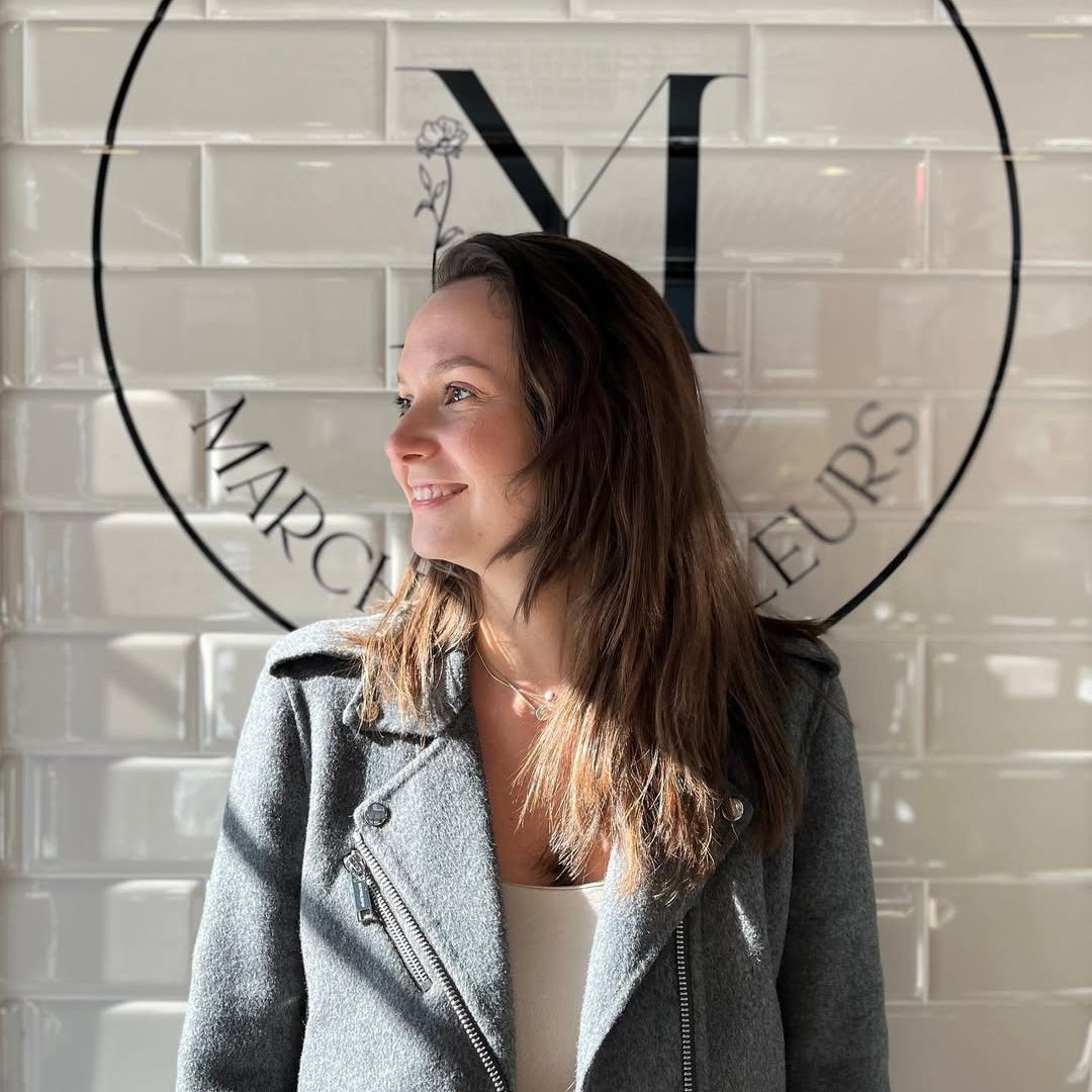 Lioudmila Zoueva, souriante devant le logo du Marché aux Fleurs, éclairée par la lumière naturelle.