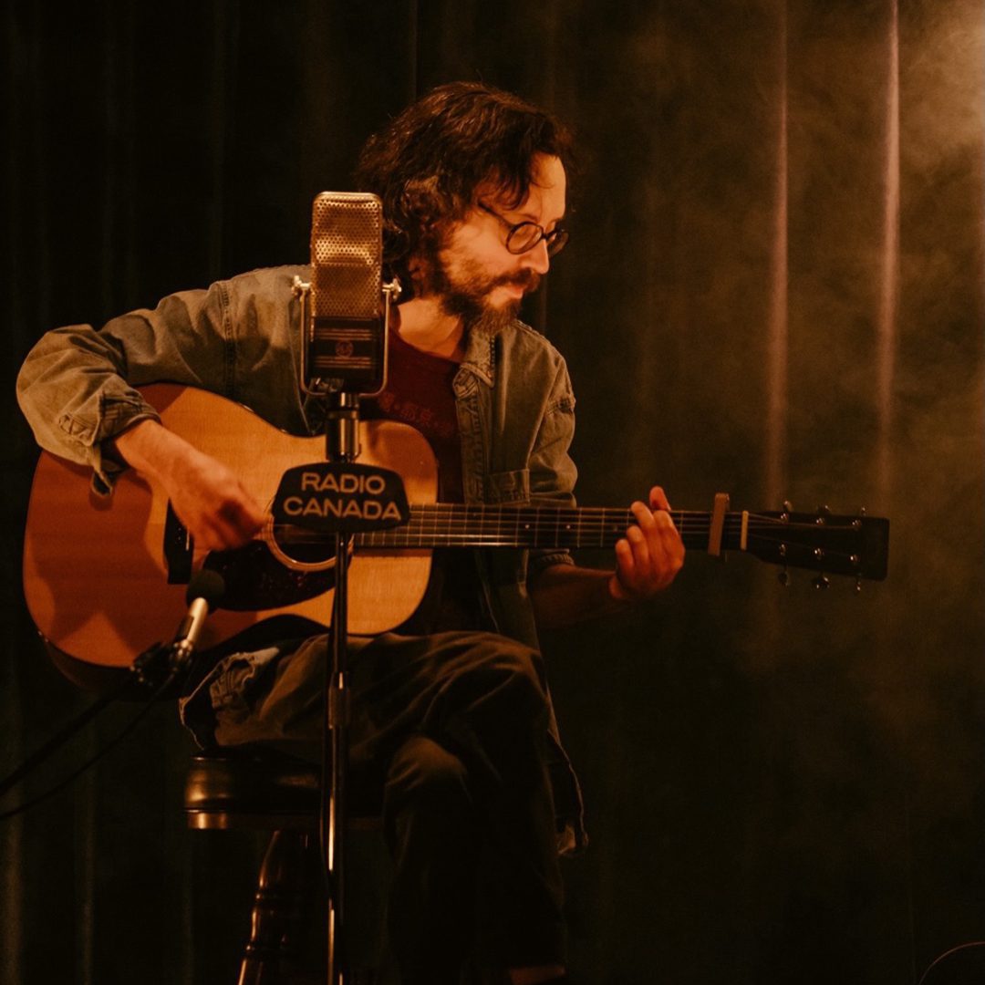 Musicien assis jouant de la guitare devant un micro dans une ambiance chaleureuse et tamisée.