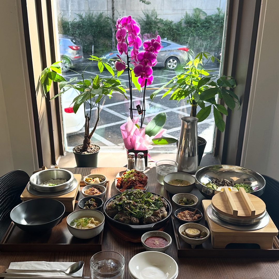 Table remplie de plats coréens variés devant une fenêtre ensoleillée avec des plantes et une orchidée rose.