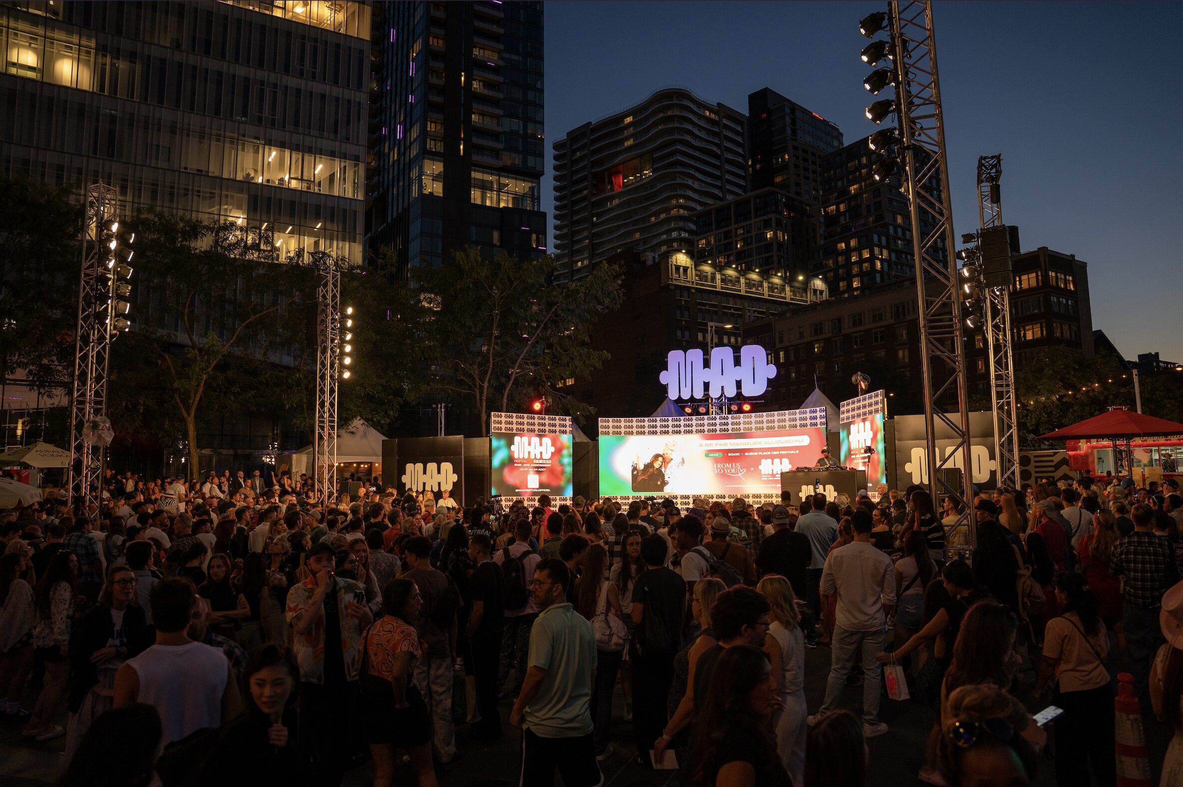 Foule rassemblée devant la scène principale du Festival M.A.D. au cœur du centre-ville en soirée.